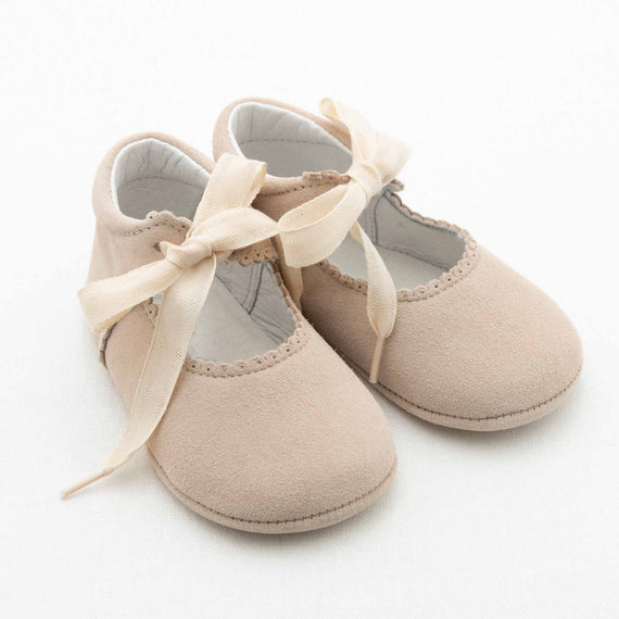 A pair of Tan Suede Tie Mary Janes from Baby Beau & Belle. These handmade baby shoes are crafted from soft suede-like material and feature delicate scalloped edges around the opening, fastened with cream-colored ribbon ties shaped into bows. The shoes are set against a plain, white background.