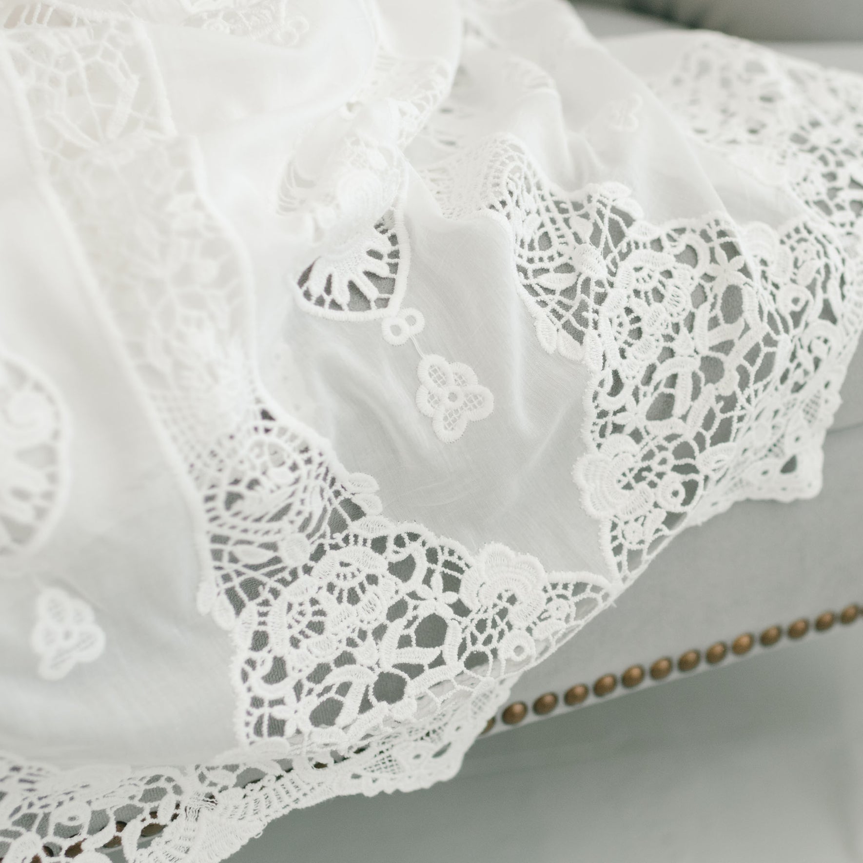 Close-up of Grace Christening Gown & Bonnet draped over a light gray piece of furniture. The lace, reminiscent of a Christening Bonnet, features intricate floral patterns and delicate detailing, adding a touch of elegance to the scene. The edge of the fabric appears to be finished with scalloped designs.