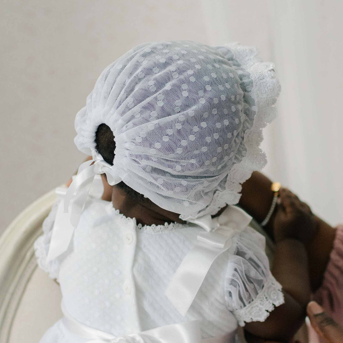 Close up of the back of the Stella Lace Bonnet as worn by a baby against a neutral background. The back details show white dotted lace and silk ribbon ties that secure the bonnet.