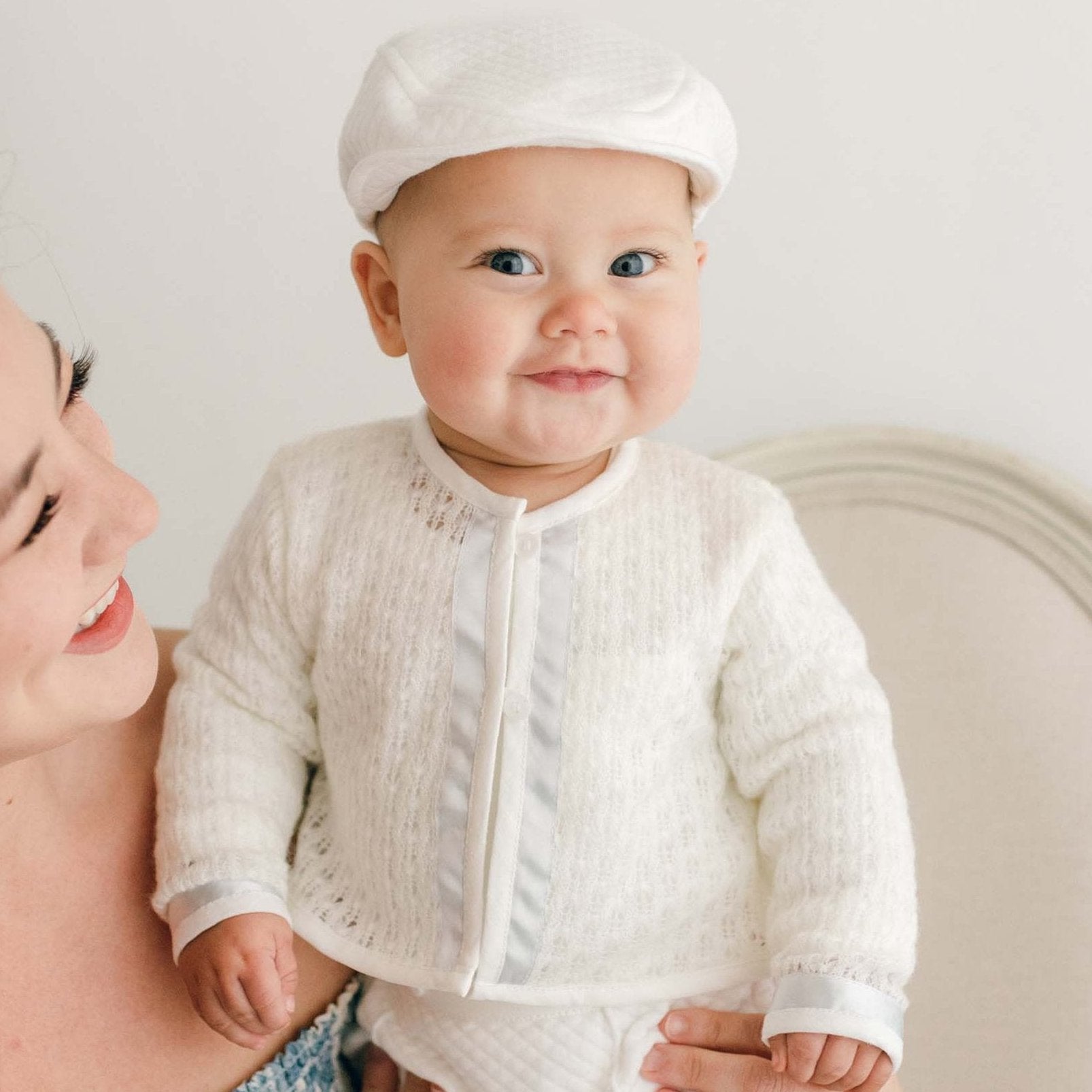 Baby boy smiling with his mother and wearing the Owen Knit Sweater made from a soft knit in ivory and trimmed with ivory linen and blue silk ribbon