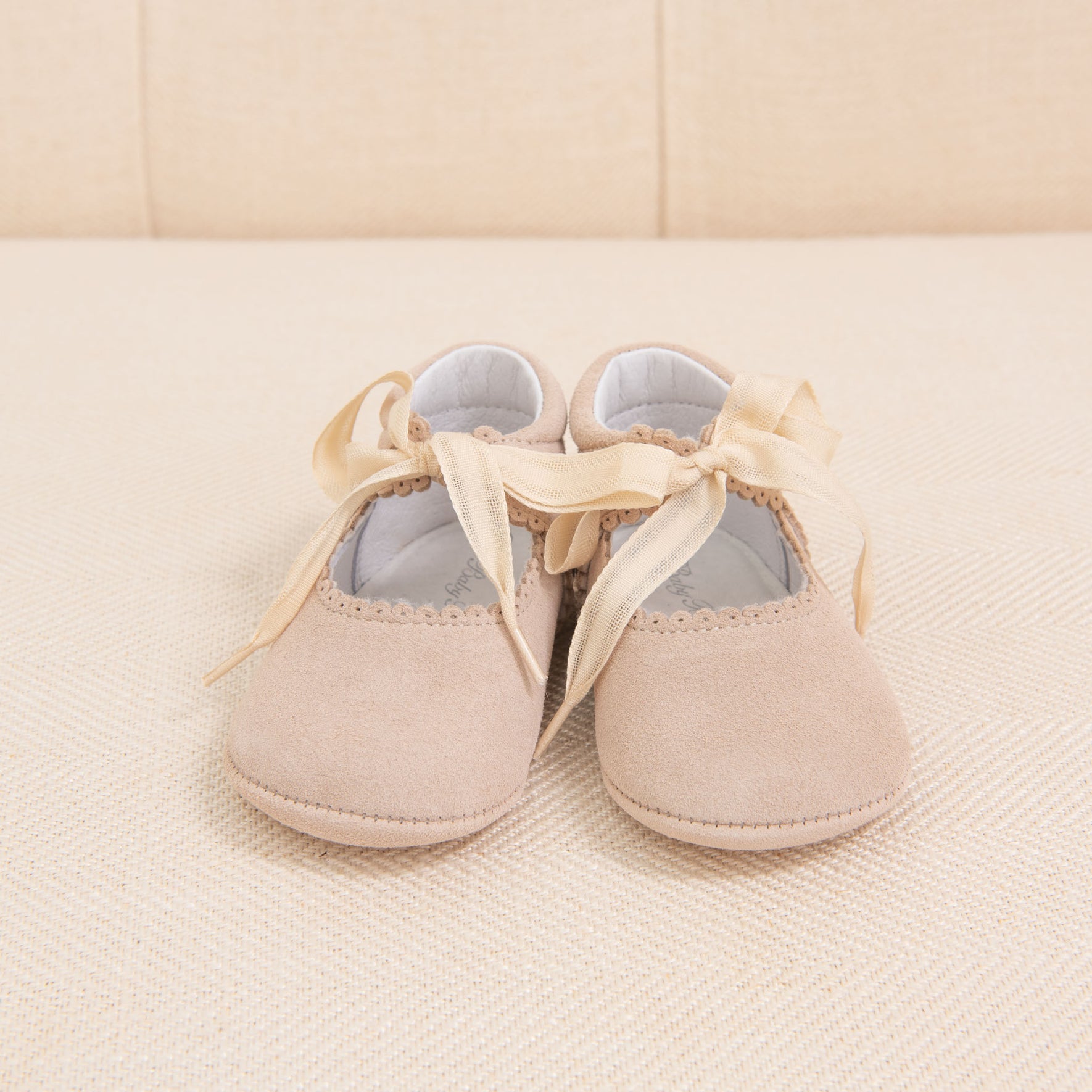 A pair of Tan Suede Tie Mary Janes by Baby Beau & Belle, crafted from soft suede leather and adorned with satin ribbons tied into bows, is placed on a cream-colored textured surface. The handmade shoes feature subtle scalloped edges along the opening.