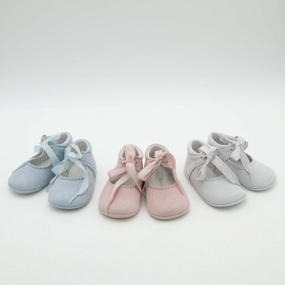 Four pairs of Thea Suede Tie Mary Janes are arranged in a row against a white background. The pastel-colored Mary Janes include two pairs in blue, one pair in pink, and one pair in light gray. Each pair features a ribbon tied in a bow on top.