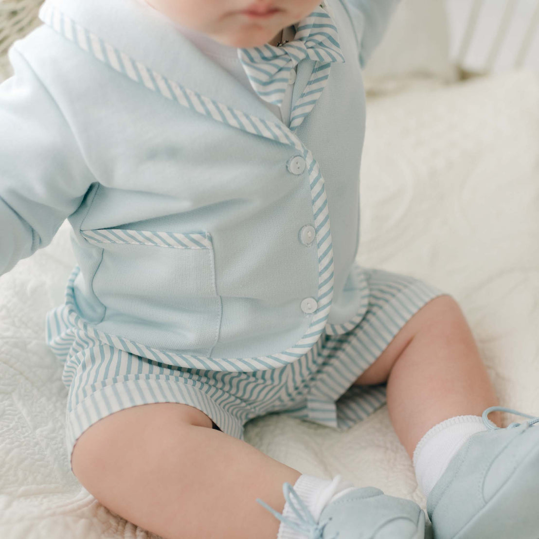 A baby wearing a Theodore Shorts Suit in light blue with striped details, including a bow tie, sits on a white quilted surface. The outfit consists of a jacket, matching shorts, socks, and soft shoes. The baby's face is not visible in the image.
