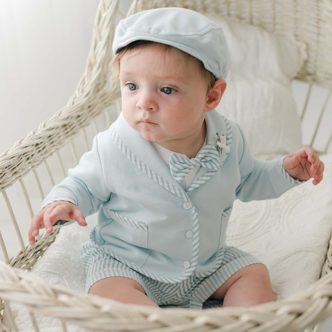 A baby dressed in the Theodore Shorts Suit, which includes a long-sleeved jacket, shorts, and a light blue striped bow tie, sits in a white wicker chair with a matching cap. Perfect for special occasion baby clothes, the background features softly lit white and neutral tones.