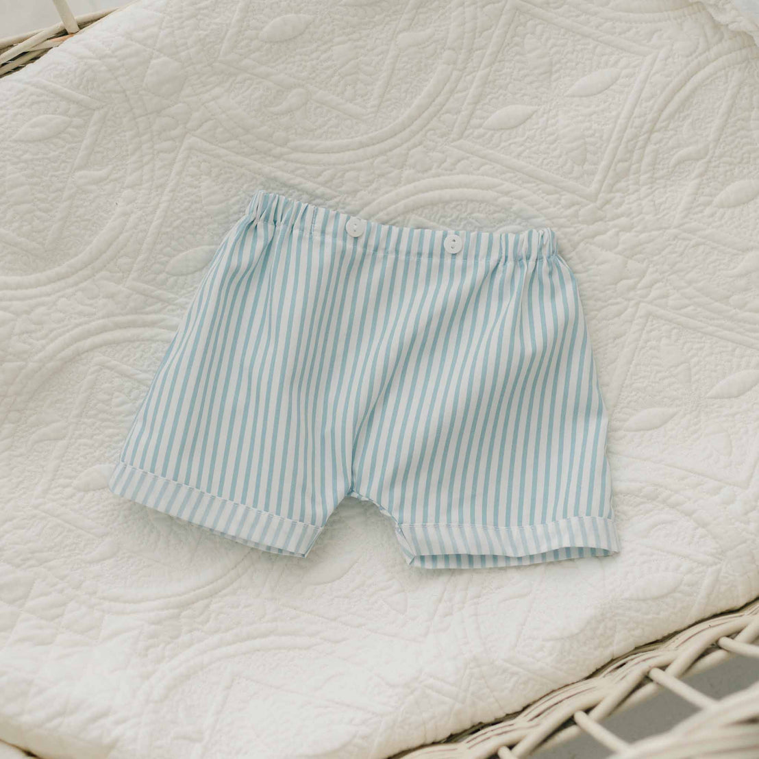 The Theodore Shorts Suit, a blue and white striped outfit designed for baby boys, features an elastic waistband and two white buttons. Ideal for special occasions, the shorts boast a neat, sewn hem and are displayed on a textured white quilt.