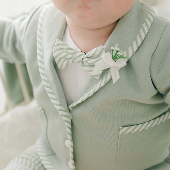 A close-up of a baby wearing the Theodore Bow Tie & Boutonniere suit. The light green suit, handmade in the USA, features white and green striped cotton trim and a matching bow tie. White buttons adorn the front, and there is a small floral boutonnière on the left lapel. The baby's face is partially visible.