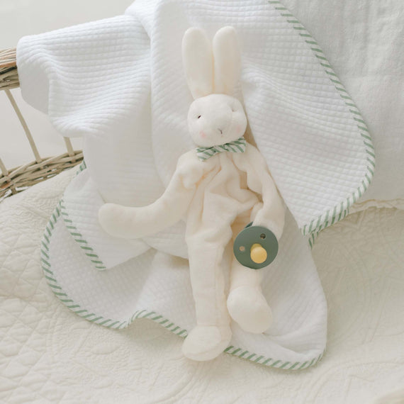 The Theodore Bunny Buddy, a white plush bunny with a pastel green bow tie, sits on a white blanket adorned with green trim. Designed with baby safety in mind, the bunny is holding a green pacifier. Placed on a wicker surface, this charming set makes for an ideal christening gift.