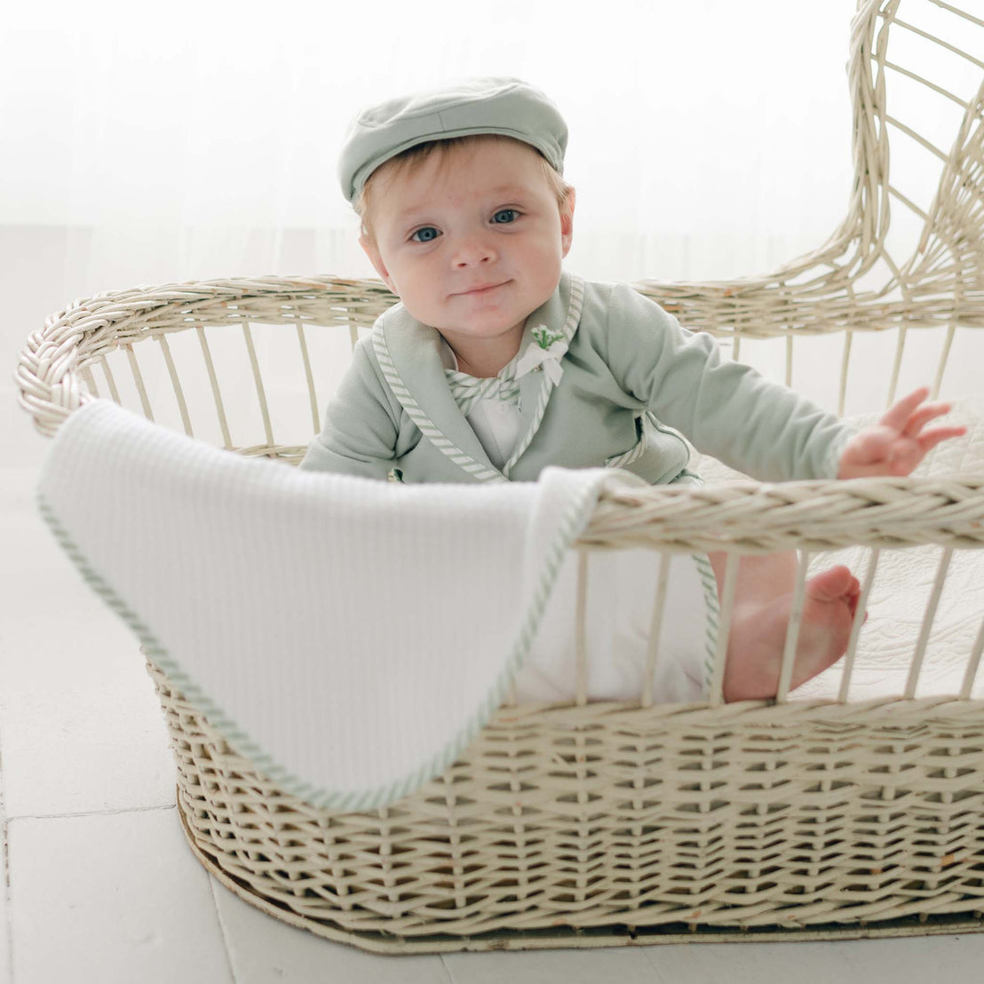 A baby dressed in the Theodore Shorts Suit in light green sits in a woven bassinet. A matching cap completes the charming ensemble, while a white cloth with green trim drapes over the side. The background is minimal and softly lit, highlighting the baby's elegant attire.