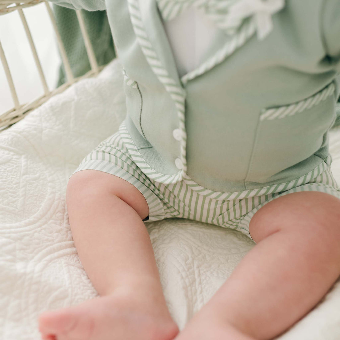 A baby is sitting on a white quilted blanket, dressed in the Theodore Shorts Suit, which features a light green outfit with white stripes. The ensemble includes an adorable jacket and shorts, resembling a handcrafted baby boy suit ideal for special occasions. The image focuses on the baby's torso and legs, leaving the face unseen.