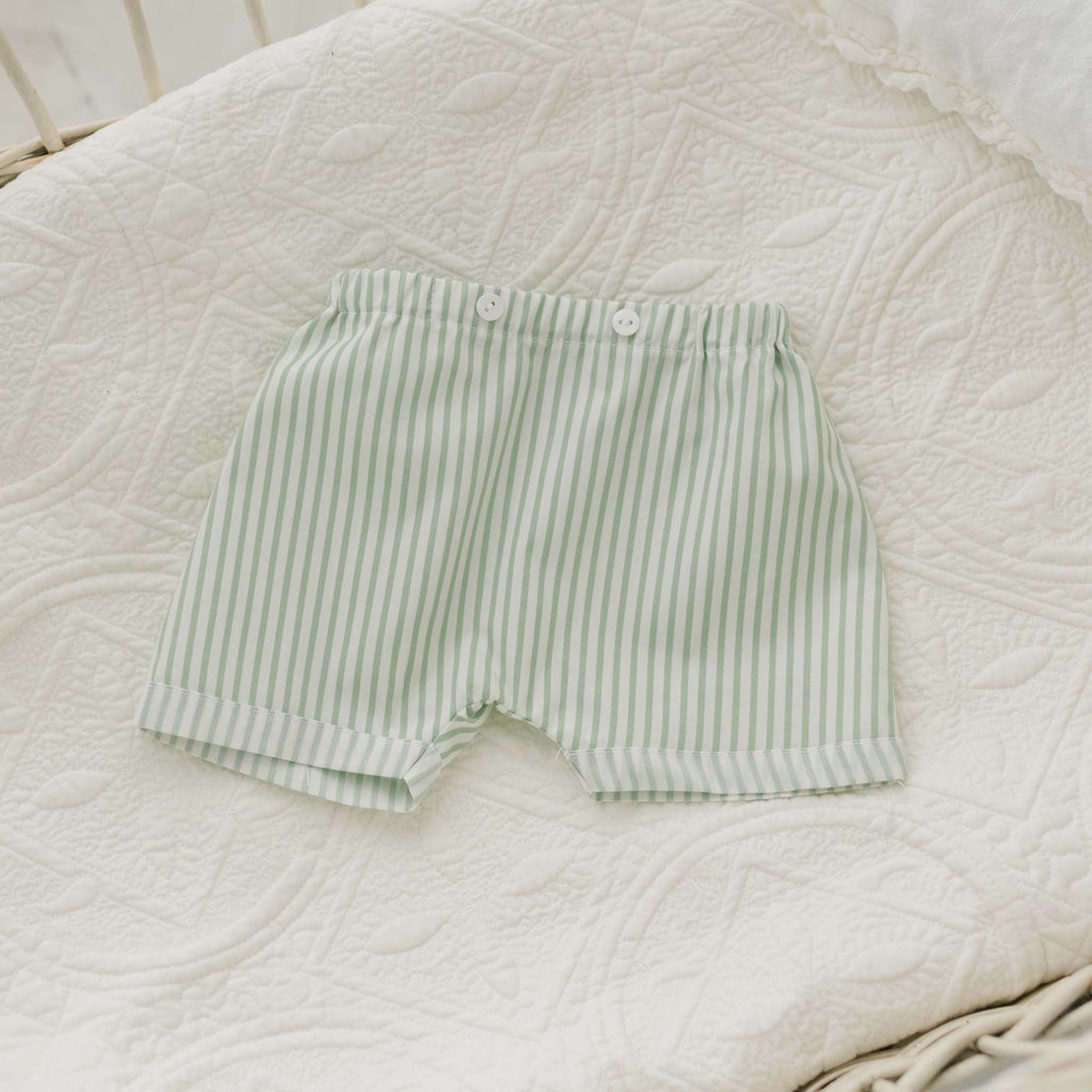 The Theodore Shorts Suit, a handmade baby boy outfit featuring light green and white striped shorts with an elastic waistband and white buttons, is displayed on a quilted, white patterned blanket. The suit includes white trim at the leg openings and is ideal for special occasions.