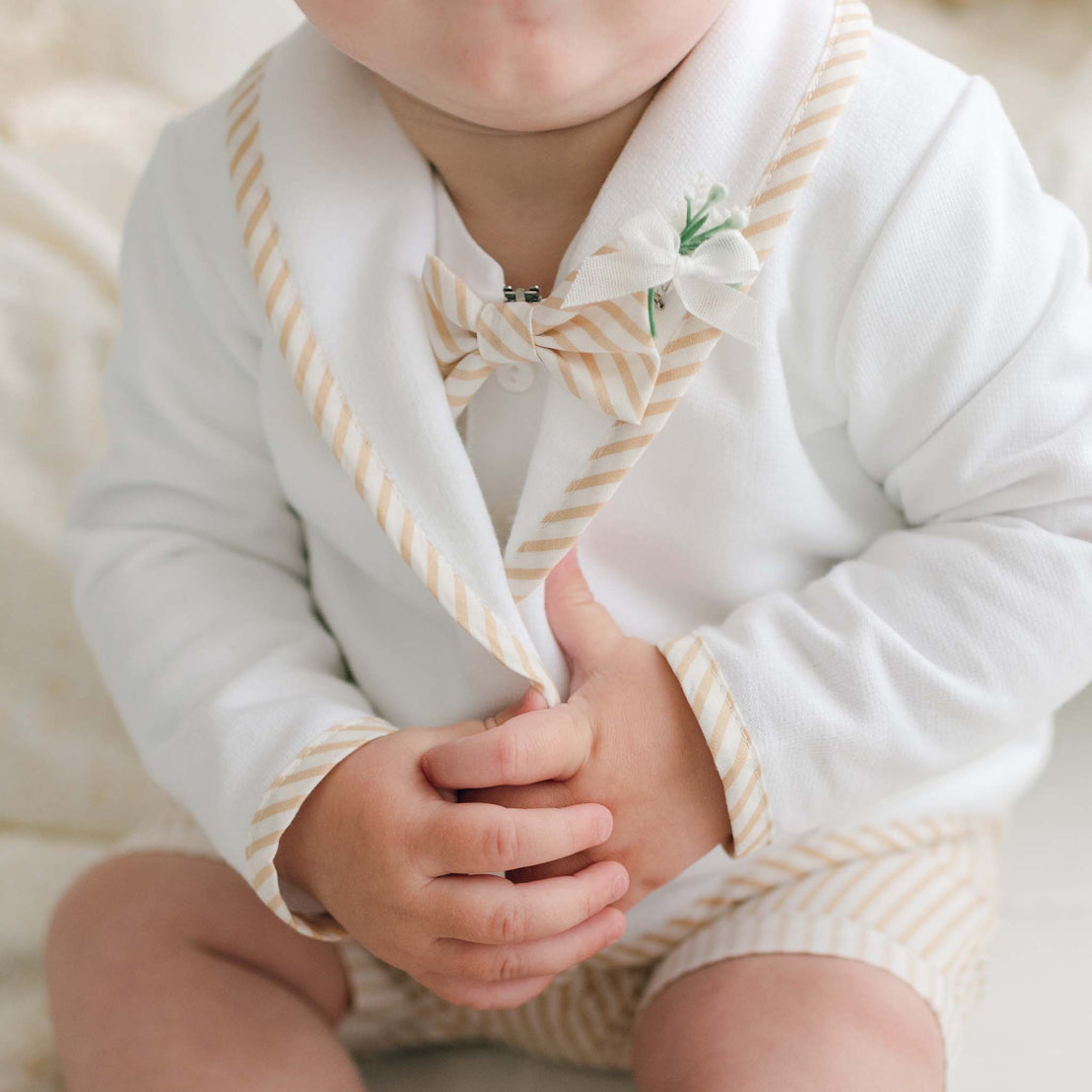 A baby wearing the Theodore Bow Tie & Boutonniere outfit, featuring a white attire with beige striped cotton accents on the collar, cuffs, and bow tie. The baby is also holding the lapels of the jacket. Handmade in the USA, with a beautifully out-of-focus background.