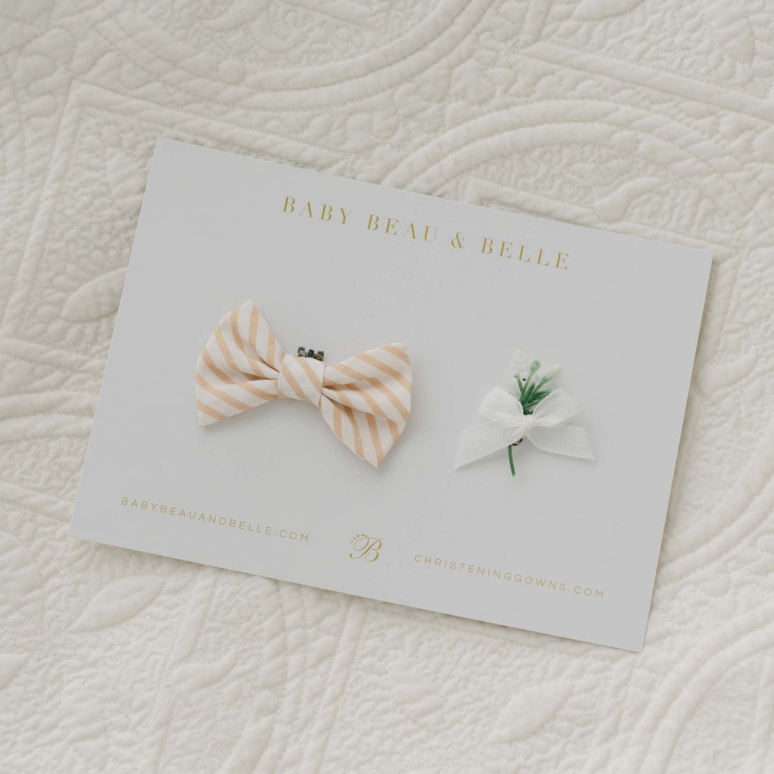 A product card featuring the Theodore Bow Tie & Boutonniere, which includes a beige and white striped cotton bow tie paired with a small white boutonniere. The card displays the brand name "Baby Beau & Belle" along with their website URLs, "babybeauandbelle.com" and "christeninggowns.com," set against a quilted fabric backdrop. Handmade in the USA.