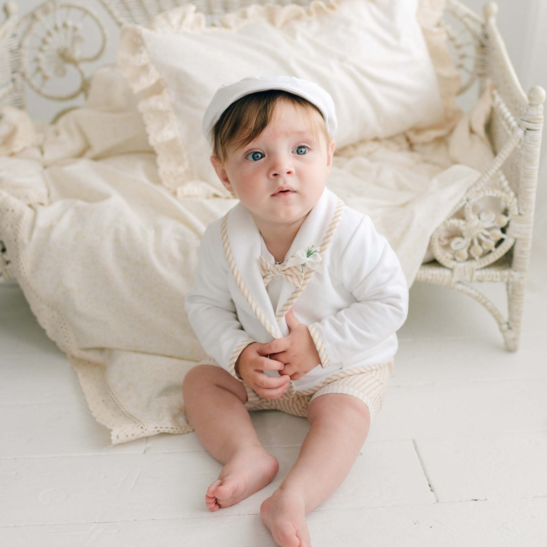 A baby wearing the Theodore Shorts Suit with beige stripes and a white cap is sitting on a white floor in front of a vintage-style bed with cream-colored blankets. The baby is looking slightly to the left with a curious expression.