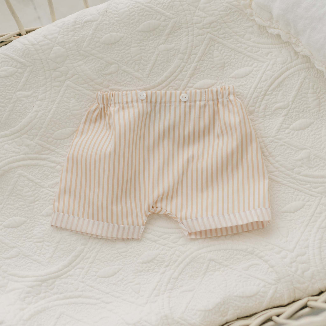 A pair of Theodore Shorts Suit, designed for baby boys with white and beige vertical stripes, lies on a textured, white quilted blanket in a wicker bassinet. Perfect for special occasion baby clothes, these shorts feature an elastic waistband and two small white buttons at the front.