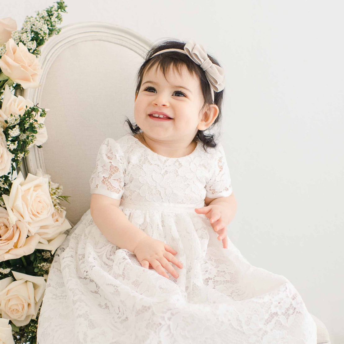 A cheerful toddler with dark hair, wearing a Rose Christening Gown & Bonnet, sits next to a chair adorned with cream roses, smiling joyfully in a light-colored room.