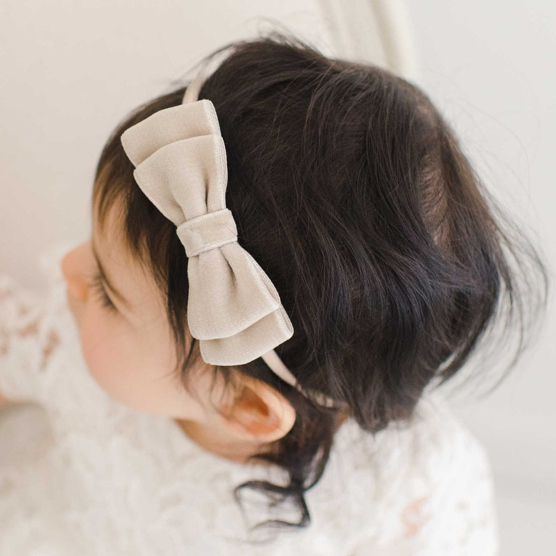 A close-up of a baby with a Rose Velvet Bow Headband in their hair, focusing on the top of their head and the light gray bow against dark hair. The visible part of their outfit suggests a