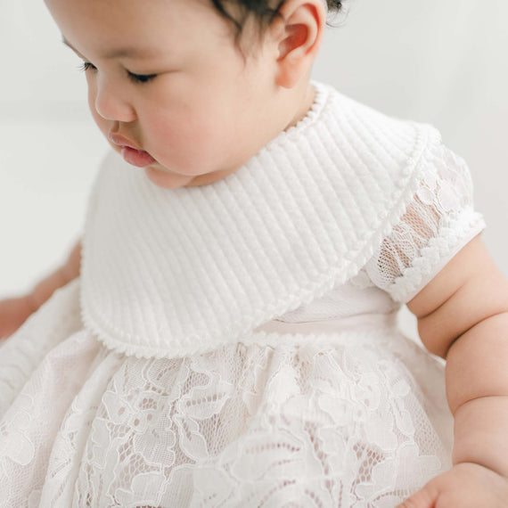 A baby wearing a Victoria Christening Bib over the Victoria Puff Sleeve Christening Dress. The photo captures the baby from the shoulders up with soft focus on the background.