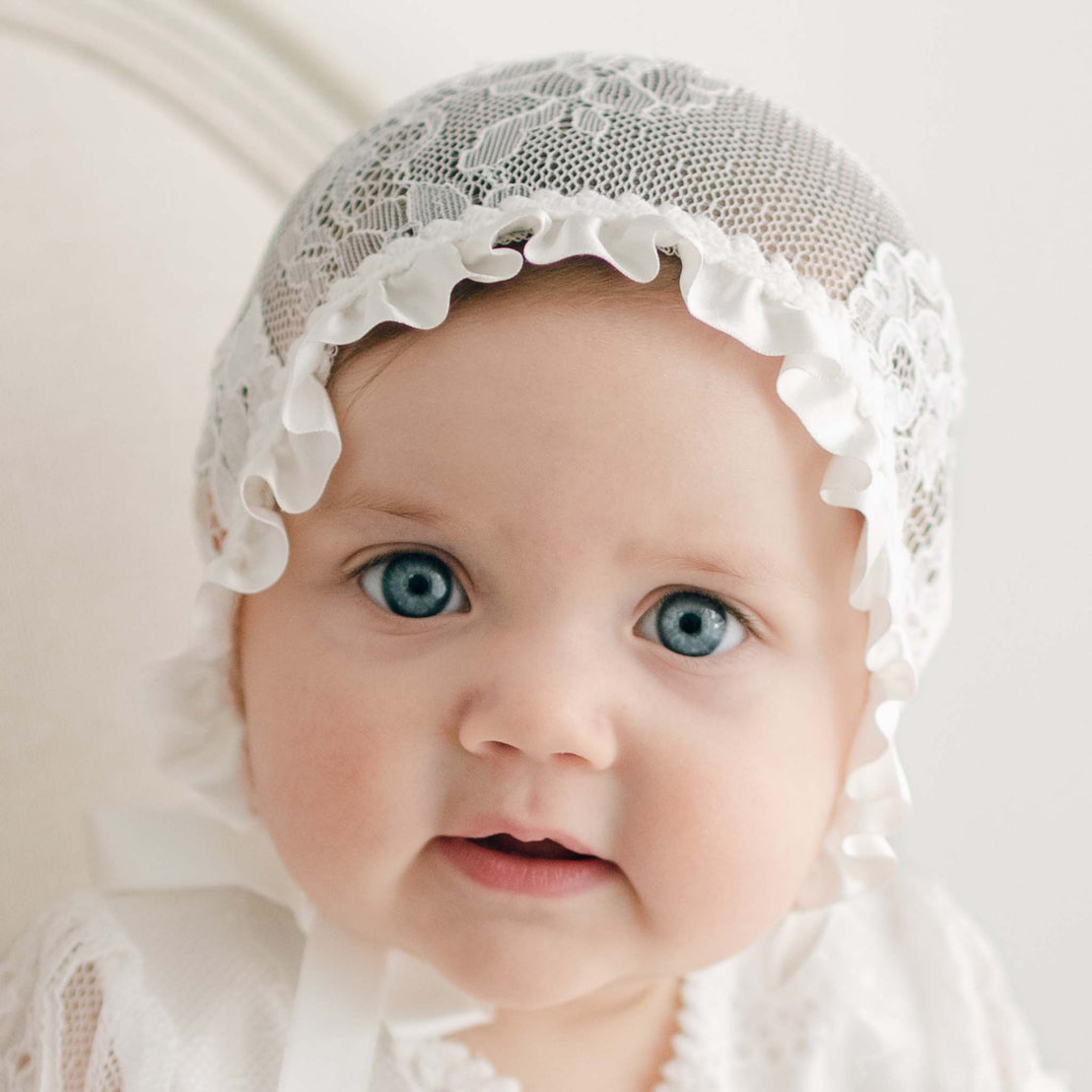 A baby with blue eyes is wearing a white Victoria Puff Sleeve Christening Gown & Bonnet, a matching outfit, and an exquisite baptism gown. The baby's expression is neutral, and the background is plain and light-colored.