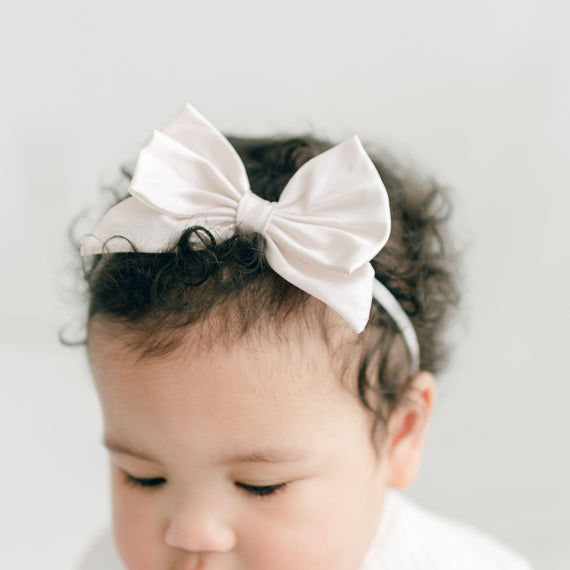 A close-up of a baby wearing a Victoria Silk Bow Headband in pink champagne. The baby has curly hair and is looking slightly downward. The background is blurred and light-colored.