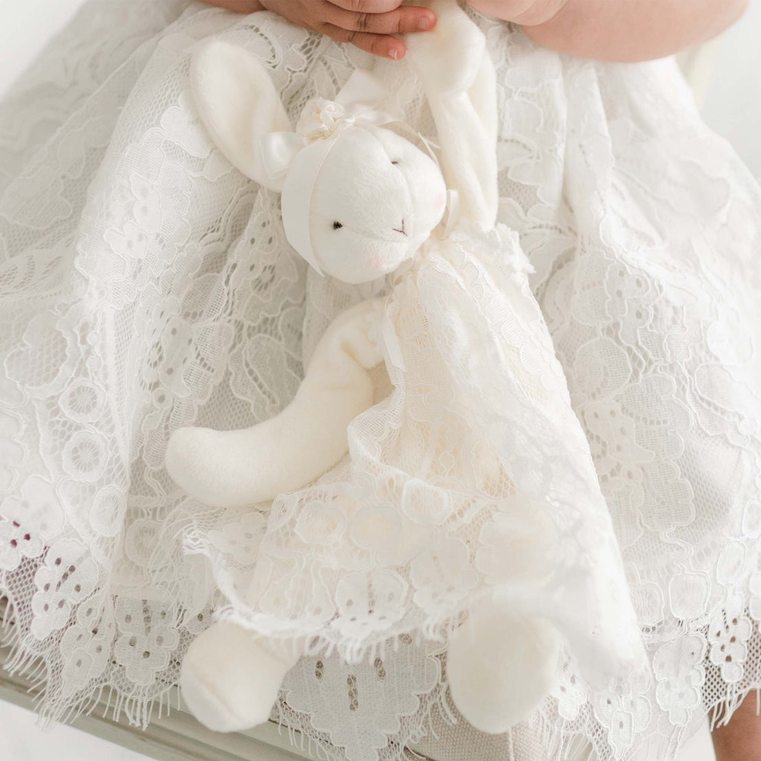 A child, dressed in a white lace dress, holds a Victoria Silly Bunny Buddy | Pacifier Holder also adorned with lace. The child's hands gently clutch the bunny's arms, which double as a baby-safe pacifier holder. The detailed lace pattern of both the dress and the toy is prominently featured in the foreground.