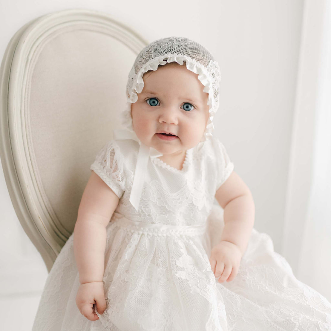 A baby sits on a beige upholstered chair, wearing a Victoria Puff Sleeve Christening Gown & Bonnet. The baby has blue eyes and is looking towards the camera. The background is softly lit and white, giving a serene and classic feel to the portrait.