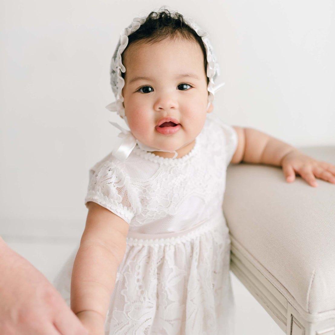 A baby wearing the Victoria Puff Sleeve Christening Dress with the Victoria Lace Christening Bonnet is holding onto the edge of a beige cushioned surface. The baby is looking towards the camera with a neutral expression, and an adult hand is gently holding one of the baby's hands.