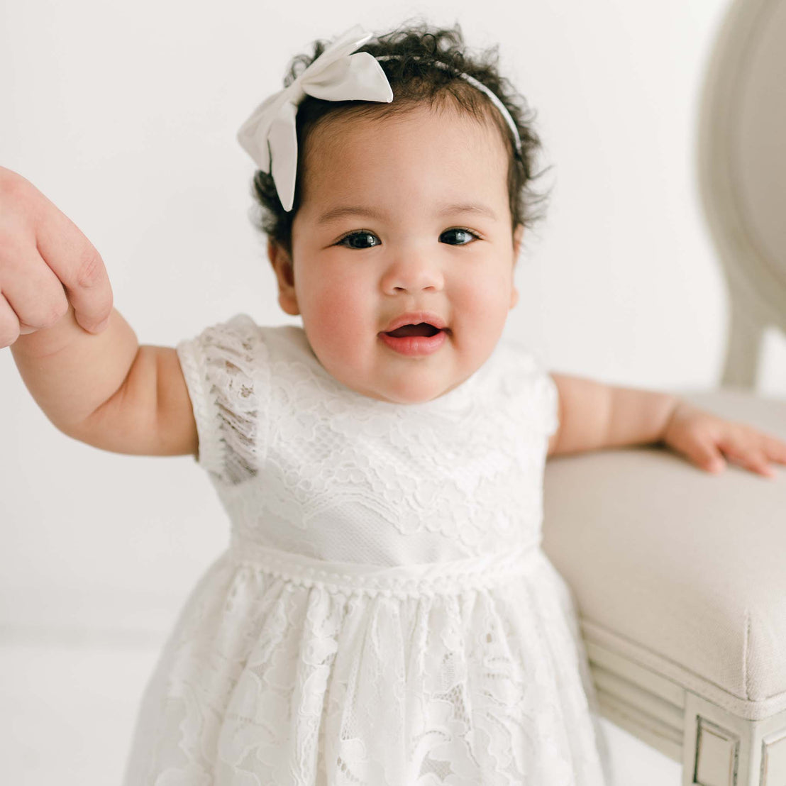 A baby wearing the Victoria Puff Sleeve Christening Dress is standing with support. The baby's right hand is holding onto an adult’s hand, while their left hand rests on a chair. The background is plain and light-colored.