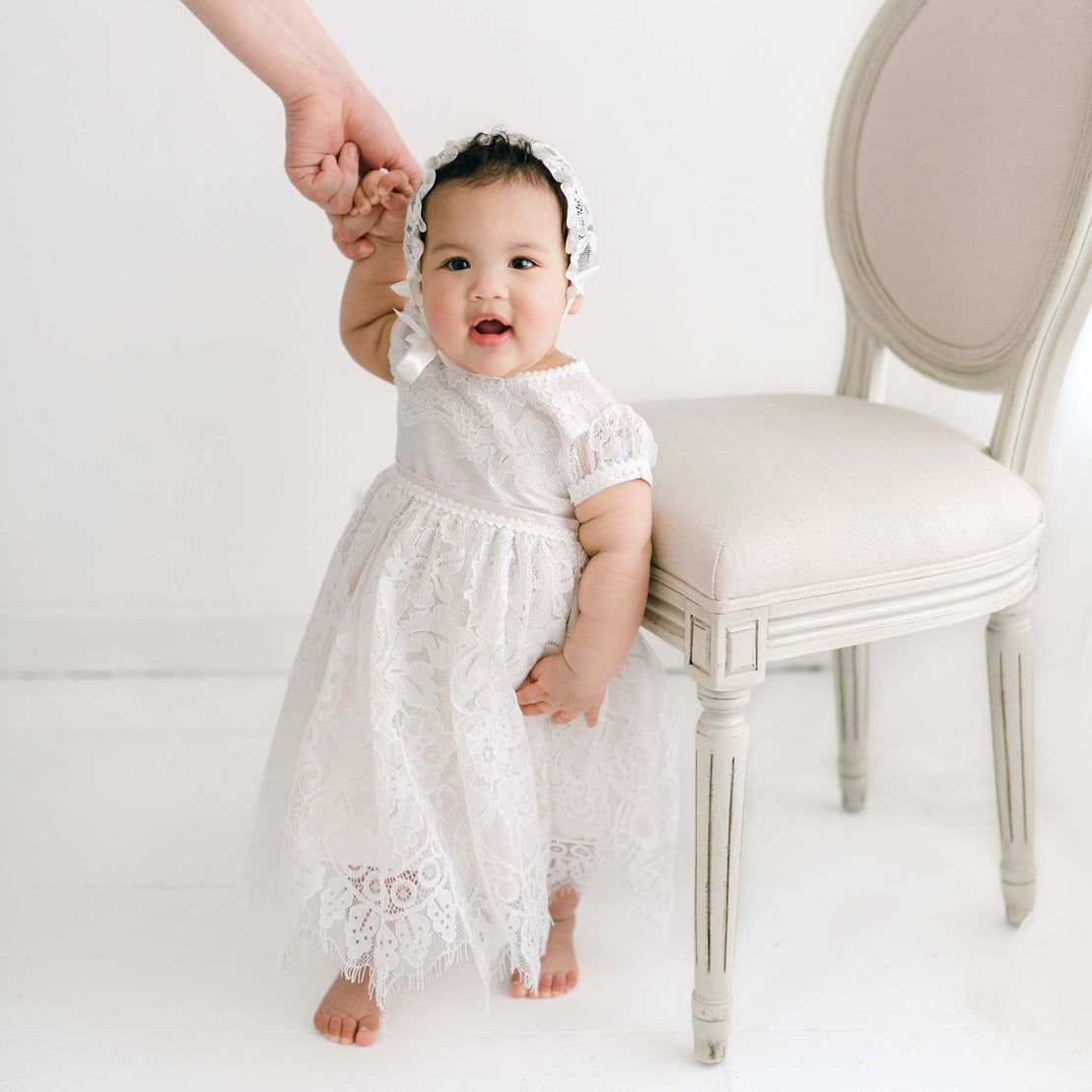 A baby wearing a Victoria Puff Sleeve Christening Dress and Victoria Lace Christening Bonnet stands next to a beige chair, holding onto an adult's hand for support. The background is minimalist with light-colored walls and flooring.