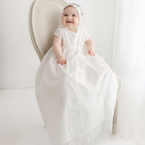 A baby is sitting on a white chair, wearing a long white Victoria Puff Sleeve Christening Gown & Bonnet with embroidered lace and a matching lace bonnet. The background is light and minimalistic with sheer white curtains. The baby is smiling and looking slightly upwards. This elegant gown, handmade in the USA, exudes timeless charm.