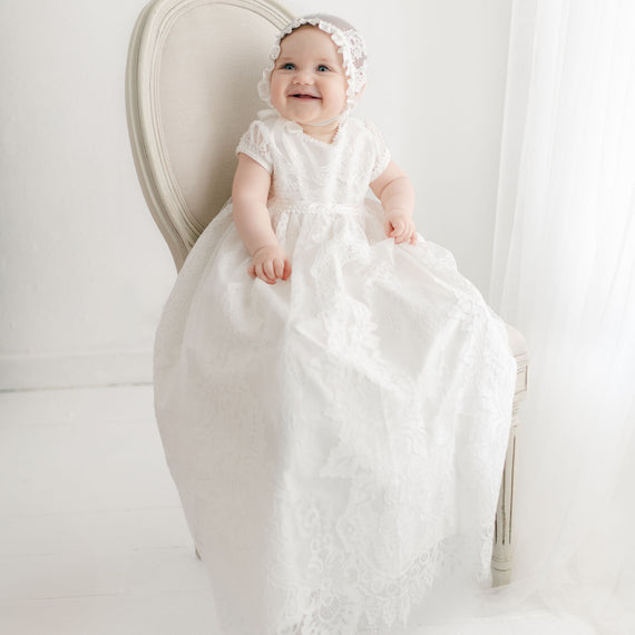 A baby is sitting on a white chair, wearing a long white Victoria Puff Sleeve Christening Gown & Bonnet with embroidered lace and a matching lace bonnet. The background is light and minimalistic with sheer white curtains. The baby is smiling and looking slightly upwards. This elegant gown, handmade in the USA, exudes timeless charm.