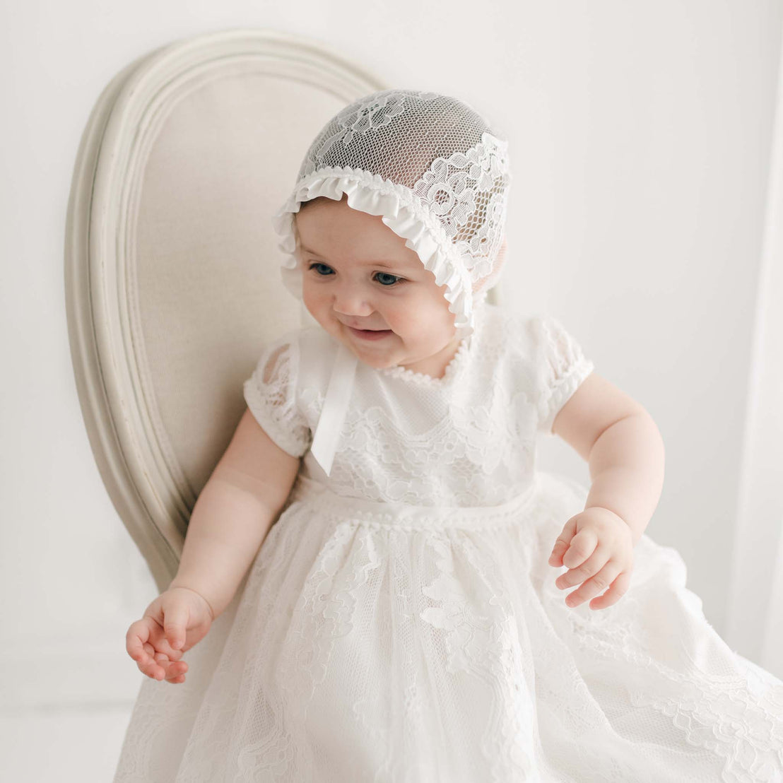 A baby wearing a white, embroidered lace Victoria Puff Sleeve Christening Gown & Bonnet sits on a cream-colored chair. The child is facing slightly to the left and appears to be smiling. The background is white and simple.