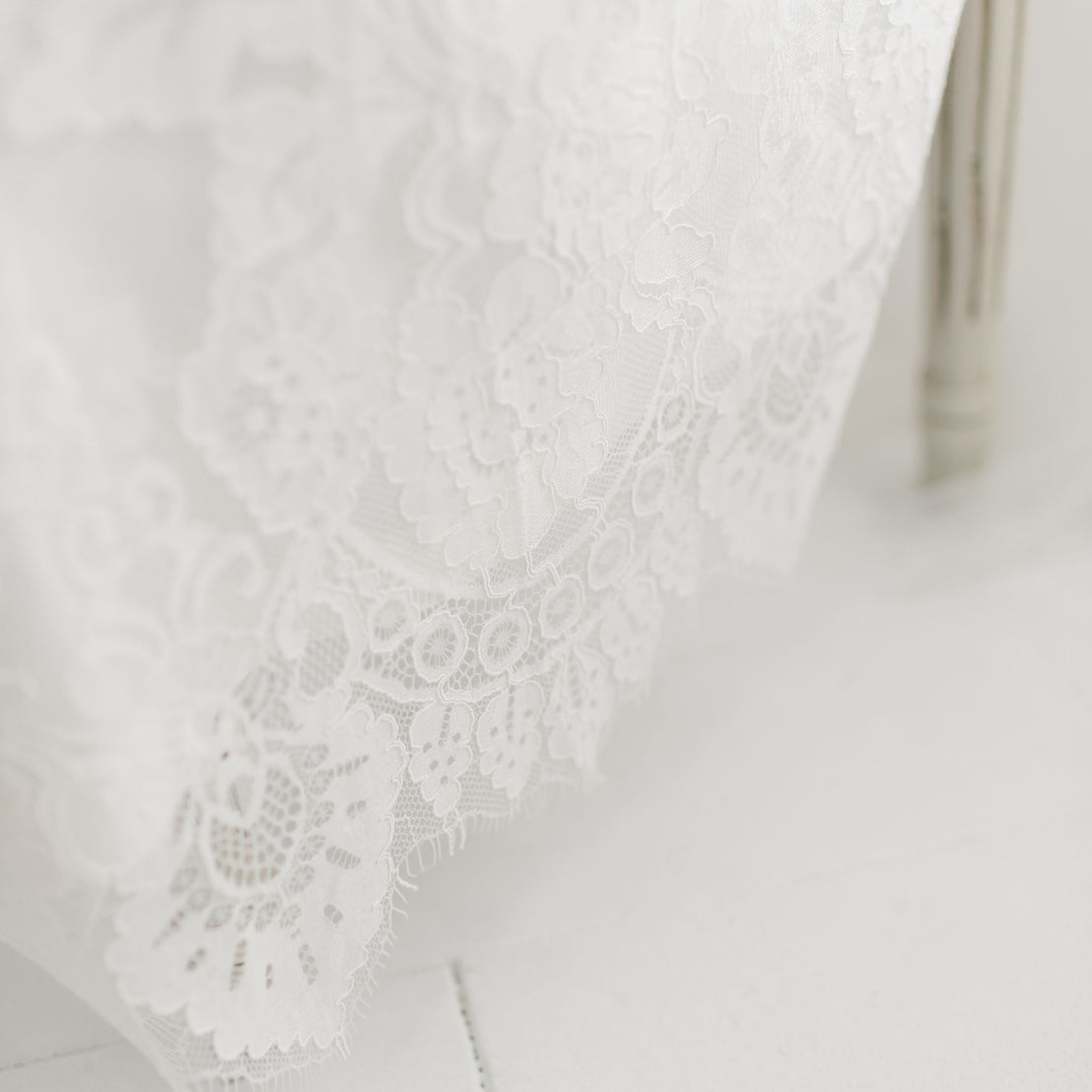 Close-up of white lace fabric with intricate floral patterns, reminiscent of a Victoria Puff Sleeve Christening Gown & Bonnet. The delicate, embroidered lace features various flower designs and fine netting, showcasing detailed craftsmanship. Part of a white chair or stool is visible to the right of the handmade USA fabric.