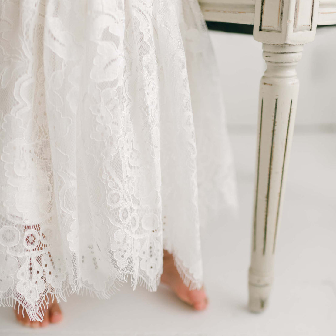 Close-up of a person standing barefoot next to a white wooden table. The person's legs are partially visible under the Victoria Puff Sleeve Christening Dress that extends to the floor. The table, reminiscent of a handmade piece, has carved vertical lines and a weathered finish.