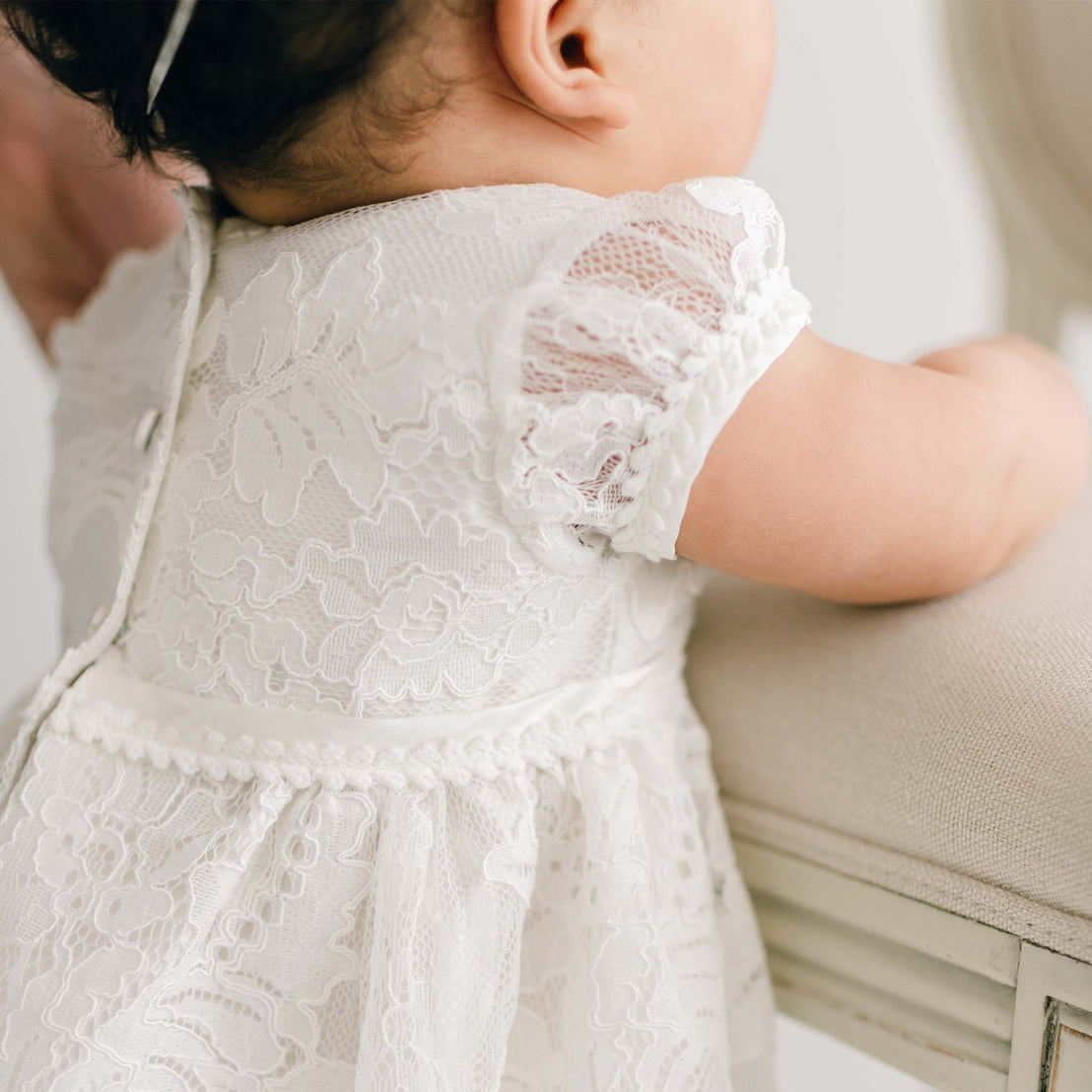 A young child wearing a white, handmade Victoria Puff Sleeve Christening Dress is seen from behind. The dress has short sleeves and button detailing along the back. The child’s dark hair is partially visible at the top of the image. The child is leaning against a light-colored surface.