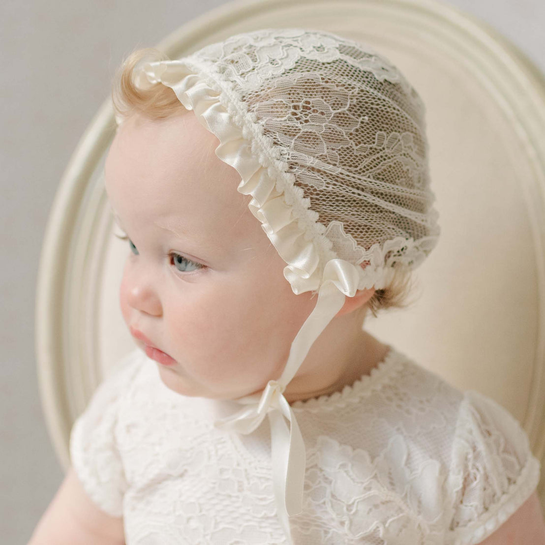 Close up photo of a baby wearing the Victoria Lace Bonnet against a neutral background.Details show the ivory floral lace and ivory silk ribbon that fits the face. Part of the Christening Gowns Baptism Gown Girl Collection.