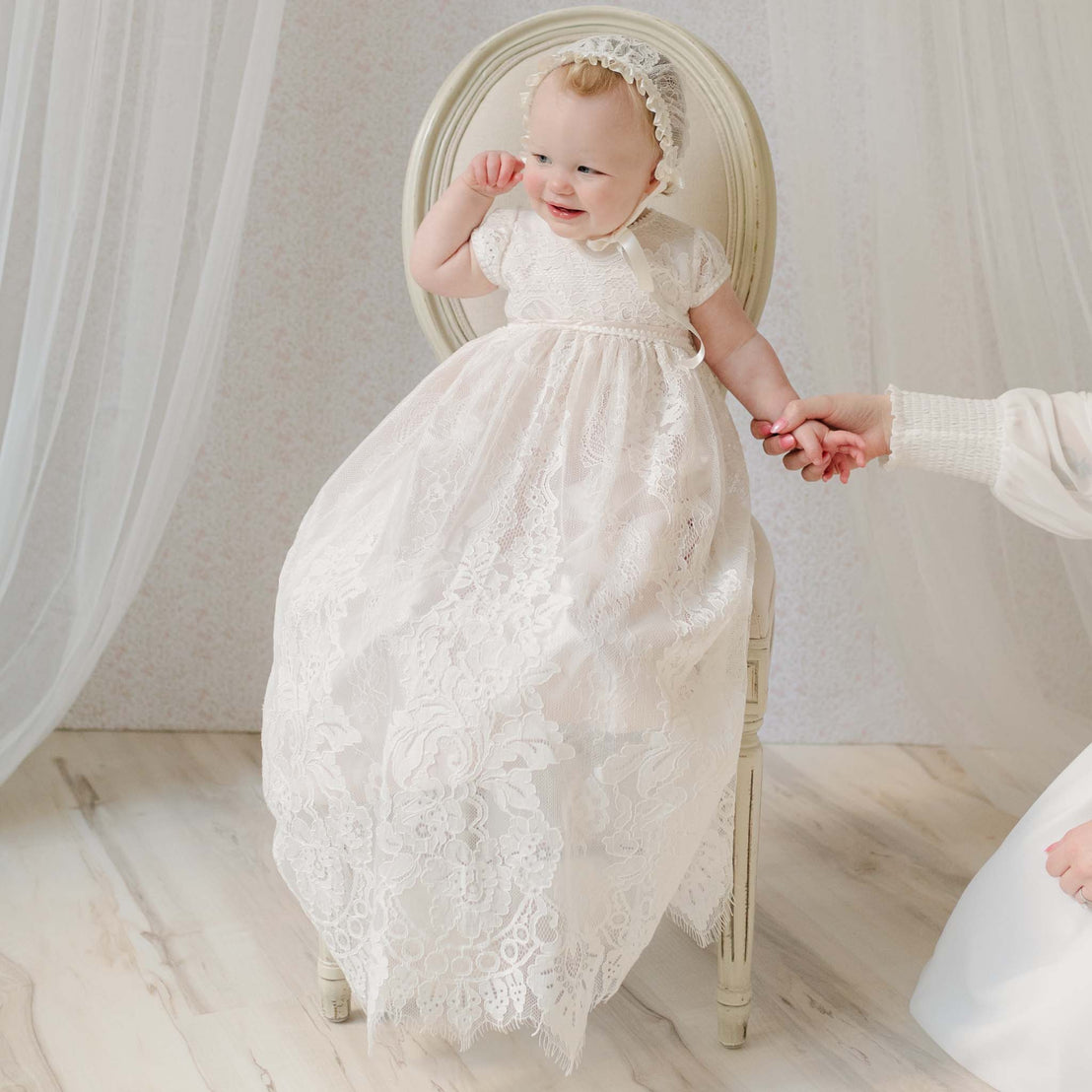Baby in the Victoria Christening Gown & Bonnet sitting on a chair and holding hands with an adult off to the side. Part of the Christening Gowns Collection.