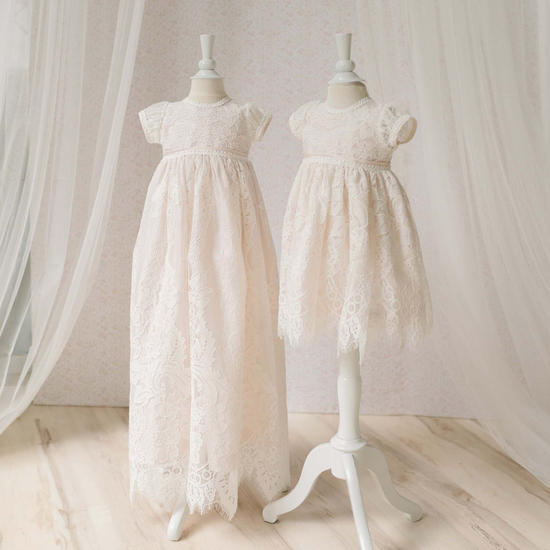 The Victoria Christening Gown and Victoria Dress on mannequins against a neutral background with sheer curtains on each side. Photo shows a difference in the length of the christening gown and baptism dress. 
