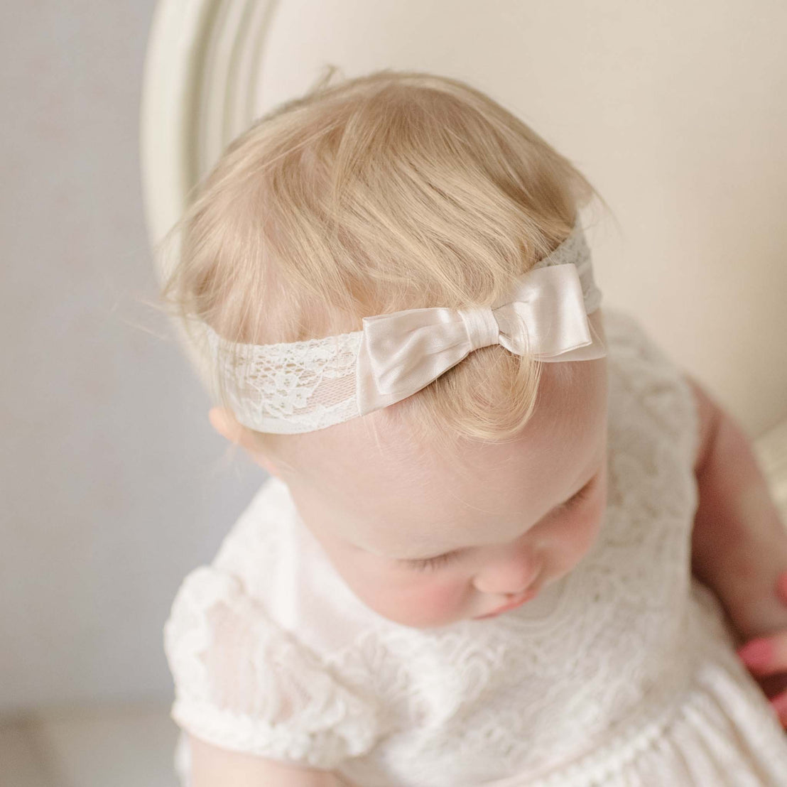 Baby wearing the Victoria Lace Headband and sitting in a chair with a blurred background. Perfect as a baptism gift.