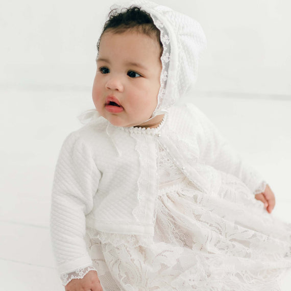 A baby wearing the Victoria Quilted Cotton Sweater over the Victoria Puff Sleeve Christening Dress and matching Lace Christening Bonnet. The sweater is adorned with pearl style buttons. The baby appears curious and is looking slightly to the side.