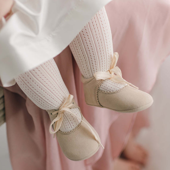 A close-up captures an infant's legs adorned in cream-colored knitted tights and beige soft shoes with satin bows. Peeking out from beneath a pink fabric, the child wears intricately crocheted Openwork Fancy Tights, set against an out-of-focus background.