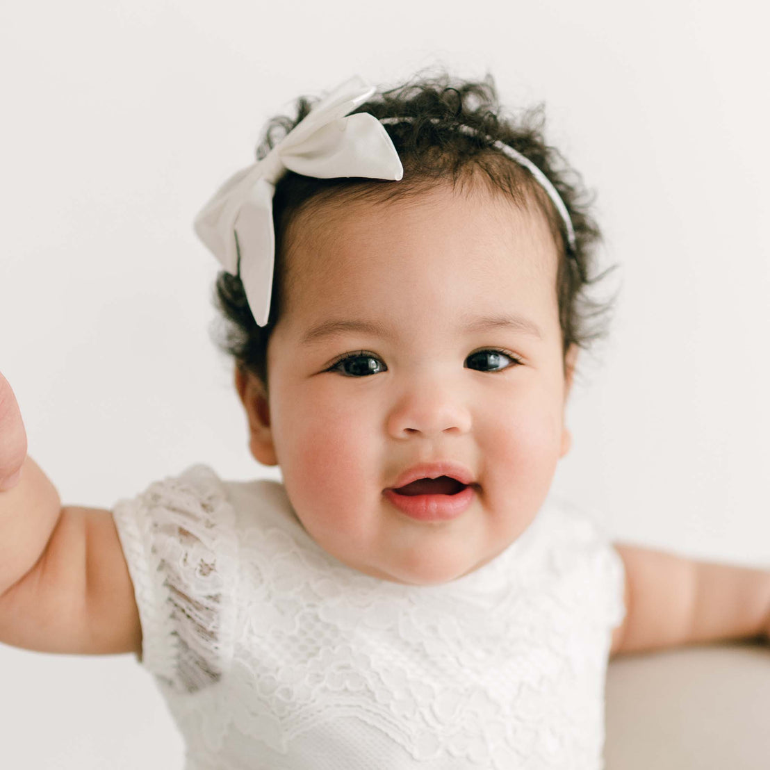 A baby girl with curly hair and a Victoria Silk Bow Headband smiles joyfully, wearing a Victoria Puff Sleeve Christening Dress against a clean, light background.
