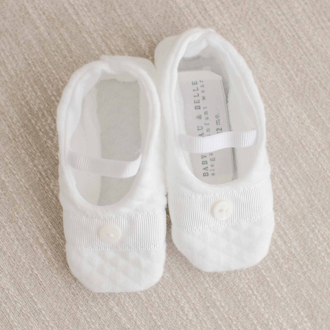 Flat lay photo of the Elijah Booties made from white textured cotton and featuring a grosgrain ribbon trim and button detail across the toe. A soft elastic strap ensures a secure and comfortable fit