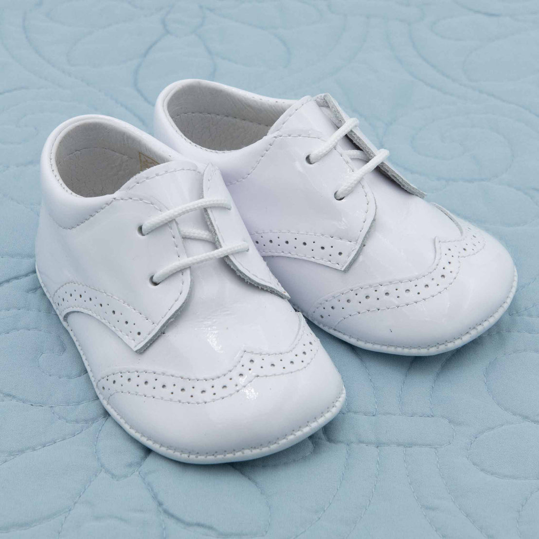 A pair of White Patent Leather Shoes placed on a light blue quilted surface. The shoes feature decorative perforations and stitching.