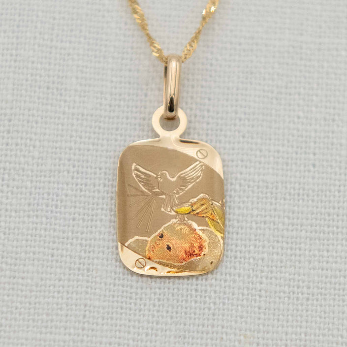A 14k gold color etched blessing charm with chain, featuring a detailed engraving of a dove soaring above a baby’s head with holy water being poured.