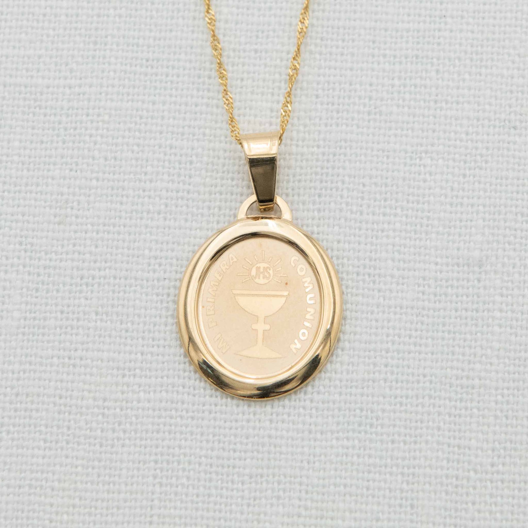 A close-up of a 14k gold oval Communion charm on a delicate gold chain, featuring an etched chalice design and "Mi Primera Comunion" inscription, displayed on a light fabric background.