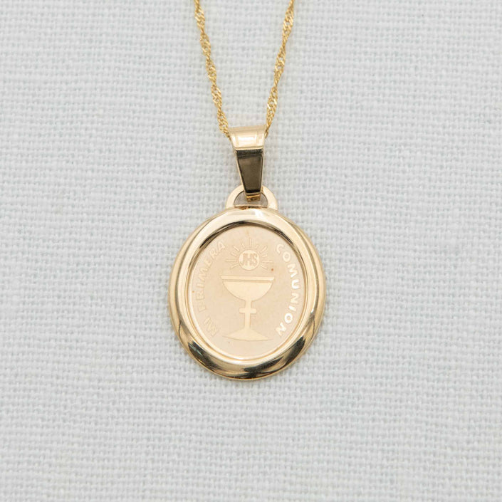 A close-up of a 14k gold oval Communion charm on a delicate gold chain, featuring an etched chalice design and "Mi Primera Comunion" inscription, displayed on a light fabric background.