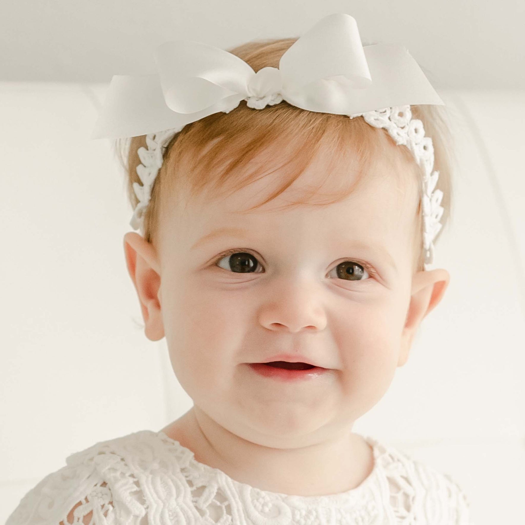A baby with light brown hair is wearing an Adeline Headband adorned with a large white bow. Dressed in a white lace Christening outfit, the baby is looking at the camera with a neutral expression. The background is plain and light-colored, making this moment feel like a precious gift.