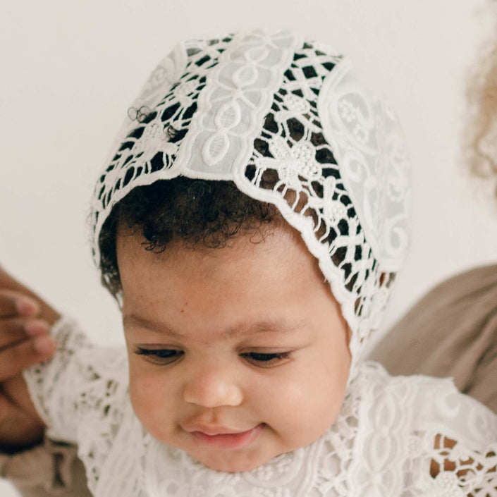 Close-up of a baby girl wearing the Adeline Lace Christening Bonnet, featuring intricate ivory lace with delicate floral detailing. The angle, as she looks down, showcases the unique openwork embroidery design.
