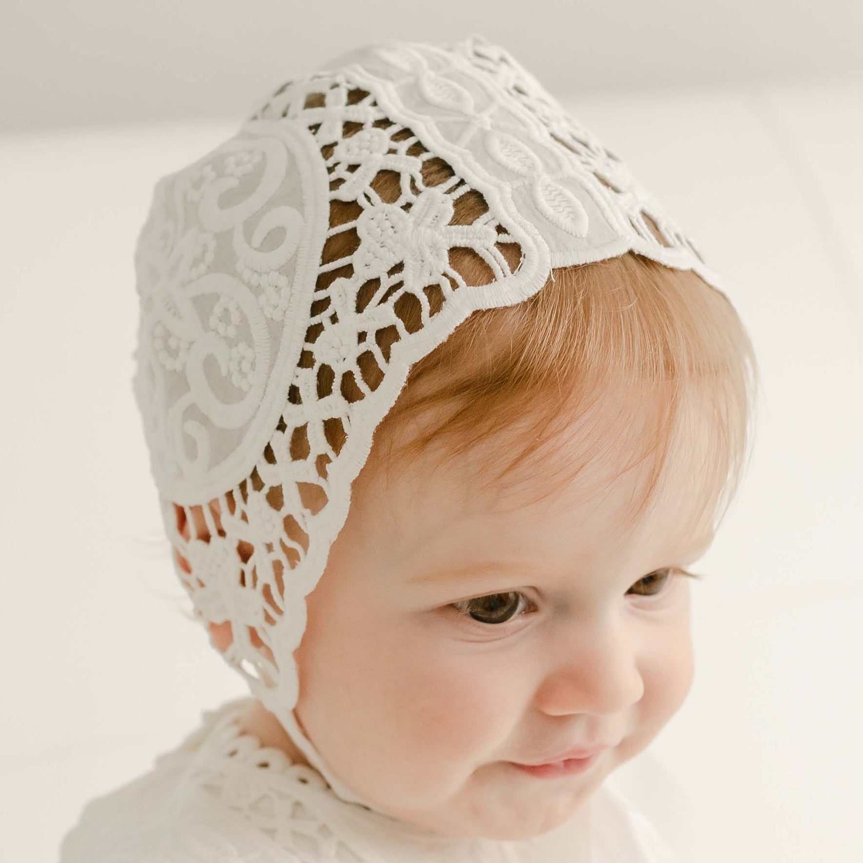 A baby girl with red hair wearing a Adeline Lace Christening Bonnet and matching lace christening dress looks downward away with a slight smile. 