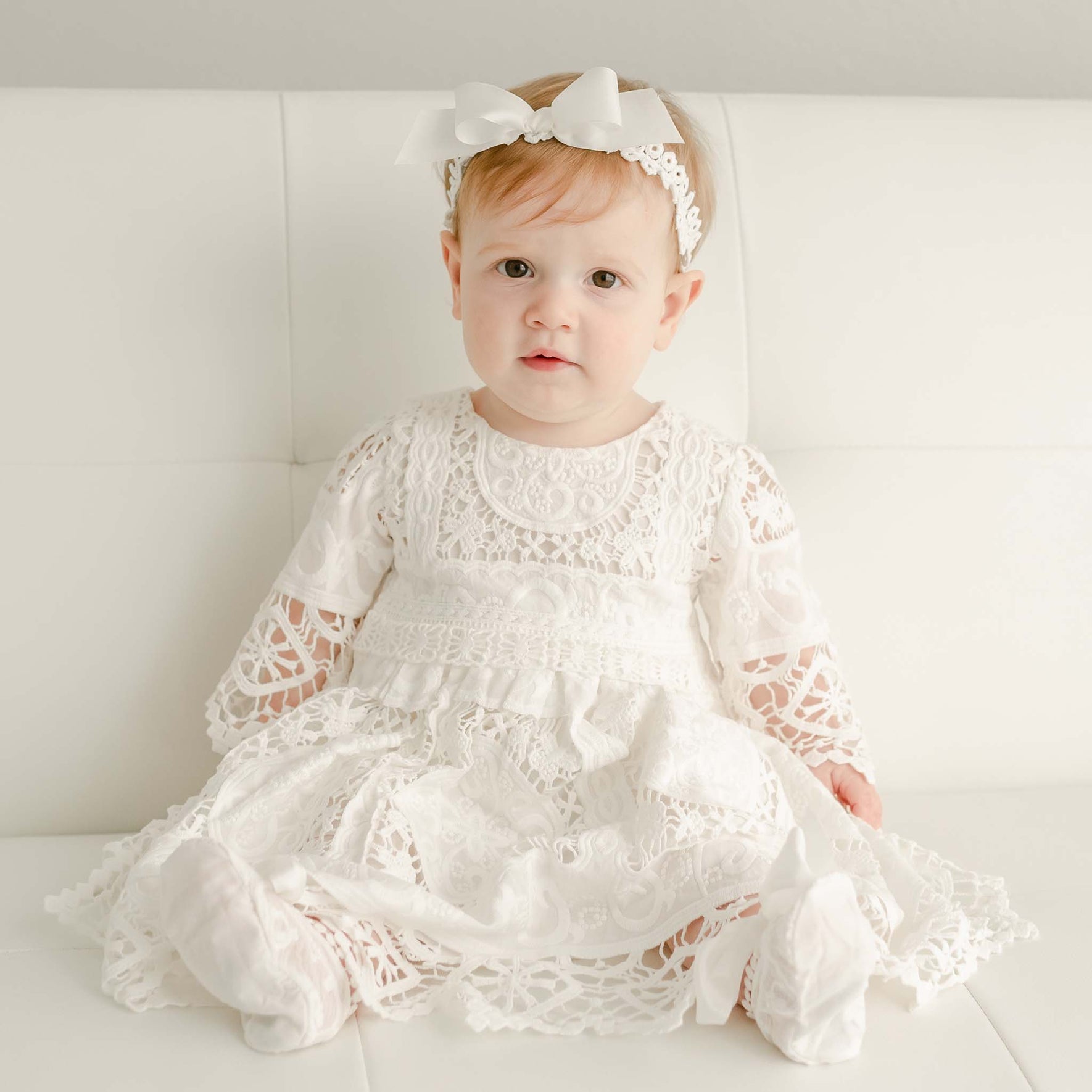 A baby is sitting on a white couch, adorned in the Adeline Lace Christening Dress & Bloomers, featuring a delicate silk ribbon sash. They are also wearing white socks and a white lace headband with a bow. The baby has light-colored hair and is looking towards the camera.